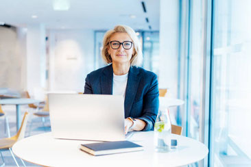 Attraktive Frau mittleren Alters mit blauem Blazer sind vor ihrem Laptop an einem runden Tisch. Blond, trägt Brille