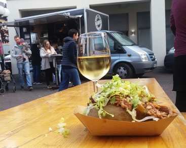 Weißweinglas und Kumpir auf Tisch. Im Hintergrund Gäste vor GOODFOOD FOOD TRUCKS an Stehtischen