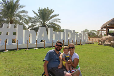 Young couple with child on grassy area in front of a sign that reads #InAbuDhabi