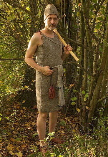 der Kund trägt einen bronzezeitlichen Trägerrock, einen genähten hohen Stoffhut und Lederschuhe, über der Schulter trägt er eine Axt