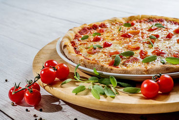Foto. Auf einem runden Holzbrett steht ein Teller mit einer Pizza. Vor und auf dem Brett liegen kleine Tomaten. 