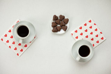 赤のハート柄のナプキンの上に置かれたコーヒーの入ったカップ＆ソーサが2客。ハート形のチョコレートが盛り付けられたお皿。