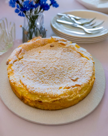 Runde Käsetorte mit Puderzucker bestäubt auf weißem Teller, im Hintergrund Tellerstapel mit Gabeln und blaue Blumen in Vase