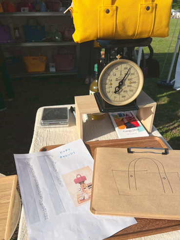 高松で夫婦で営む帆布バッグのお店Cargo Shipではクラフトイベントの時、とても軽くて使いやすいボストンバッグ”short ducks"の中に日用品を入れてピッタリ９００gになったらプレゼントがもらえるゲームを開催していて、その黄色いボストンバッグが黒い秤の上に乗り、奥には帆布バッグが並んでいます。