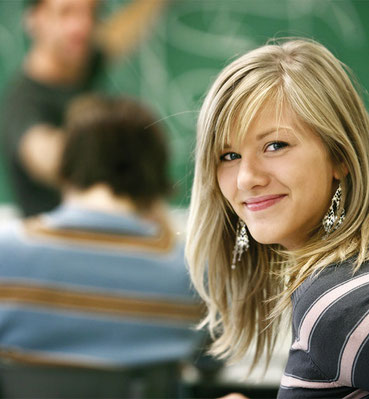 Blonde Schülerin mit langen Haaren und gestreiftem Pullover sitzt in einem Klassenzimmer mit grüner Tafel im Hintergrund