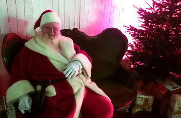 Der Weihnachtsmann sitzt neben einem Tannenbaum und vielen Geschenken.