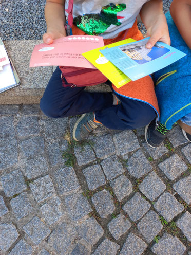 Zwei Kinder sitzen nebeneinander auf einer Mauer und halten drei Karten in der Hand. Eine gelbe, blaue und rosafarbene. Die Köpfe der Kinder sind nicht zu erkennen.