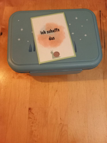 Hellblaue Brotdose steht auf Holztisch, auf ihr liegt eine Affirmationskarte mit einer kleinen Schnecke drauf und dem Spruch ich schaffe das