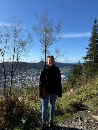 Luisa steht auf einer Lichtung auf einem Berg, sie lächelt in die Kamera. Im Hintergrund sind die Umrisse einer Ortschaft zu erkennen.