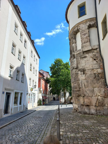 Blick in eine menschenleere Gasse von Regensburg, rechts befindet sich ein historisches Gebäude mit einem Baum, links Läden. 