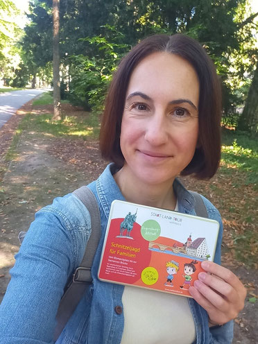 Michaela Dauscher lächelt in die Kamera und hält die Box der Schnitzeljagd in der Hand. Sie trägt ein blaues Jeanshemd und hat einen Rucksack um, die braunen Haare sind schulterlang. Im Hintergrund ein Weg und Bäume.