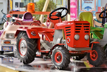 Ein leeres Kinderkarussell, im Vordergrund ein roter Trecker, dahinter andere Karussellfahrzeuge.
