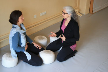 Eine Frau erklärt einer anderen Frau etwas über Energiearbeit, beide sitzen auf Yogakissen auf dem Boden in einem Yogastudio oder einem Coachingstudio
