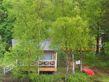 Sehenswürdigkeiten Inari: Ein gelb gestrichenes Wohnhaus steht am nahen Ufer des Inarijärvi, umgeben von Kiefern und Birken. Unweit steht ein zum Haus gehörendes Bootshaus. Ein rotes Ruderboot liegt neben dem Bootshaus.