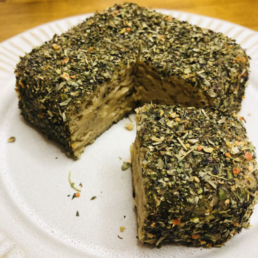 Quadratisches Stück Käse, ummantelt von grünen getrockneten Kräutern. Es ist ein Stück herausgeschnitten, sodass man das helle, weiche innere sieht.