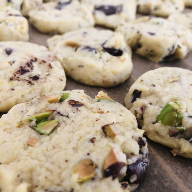 Helle Beige Cookies mit grünen Pistazienstücken und brauner leicht angeschwollener Schokoladen. Es liegen mehrere Plätzchen nebeneinander auf einem braunem Backpapier.