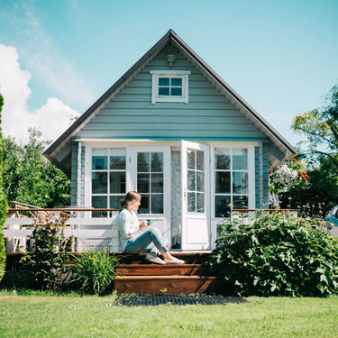Kleines Ferienhaus mit Garten, Frau sitzt auf der Veranda. Foto: Birgit Loit auf Unsplash.