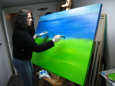 Miriam Pietrangeli steht vor ihrer Staffelei und malt ein blau-grünes Landschaftsbild auf Leinwand, das sie "Orizzonte" nennt