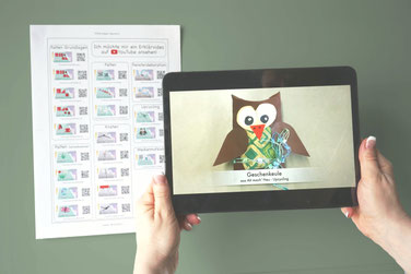 Im Vordergrund Hände, die ein Tablet halten auf dem ein Erklärvideo läuft. Im Hintergrund ist eine Videoliste mit QR-Codes auf einer Schultafel aufgehängt.