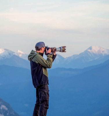 Fotograf Christopher Rau beim fotografieren mit einer Kamera, im Hintergrund sind schneebedeckte Gipfel der Alpen zu erkennen