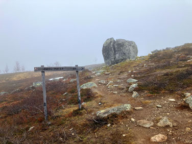 Sehenswürdigkeiten Kilpisjärvi - Dreiländereck: Ein Holzwegweiser zeigt in schwarzer Schrift den Weg zu «Pikku Malla» an. Im Hintergrund ist ein großes Felsstück, dass in der Mitte gespalten wurde.
