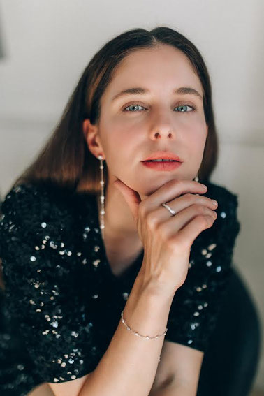 Elegante Frau in schwarzer, glitzernder Kleidung stützt ihr Kinn auf die Hand. Feiner Silberschmuck mit Ring, Armband und langen Ohrringen im Fokus bei sanftem Licht.