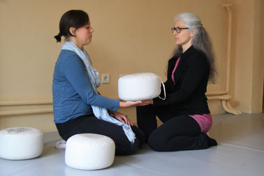 ein Yogakissen wird von einer Frau zu einer anderen Frau übergeben, beide sitzen auf dem Boden in einem Yogastudio oder Coachingstudio
