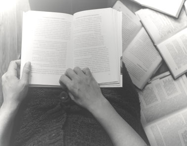 Hände halten ein offenes Buch, umgeben von weiteren Büchern – Symbol für analytische Textarbeit, Lesen und geisteswissenschaftliche Herangehensweise.