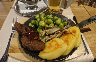 Eiserne Pfanne mit zwei Frikadellen, Rosenkohl, Pilzen und Kartoffelplätzchen. Im Hintergrund sieht man ein Glas mit Apfelschorle und ein Soßenkännchen auf einem Unterteller, beides aus Edelstahl.