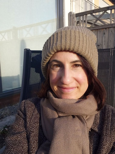 Michaela Dauscher lächelt in die Kamera, sie sitzt auf einem Stuhl im Garten, ihr Gesicht wird von der Sonne angeschienen. Sie trägt einen Mantel, eine beige Mütze und einen Schal.