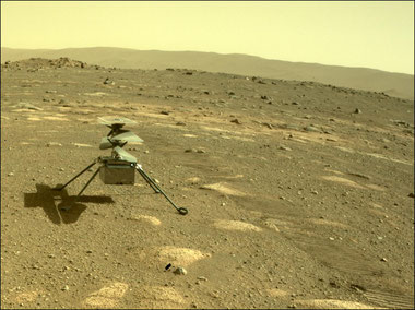 Der Mars-Helikopter "Ingenuity" auf einer sandigen, gelblich-ockerfarbenen Ebene, auf der einige Steine liegen. Im Hintergrund sieht man einen Höhenzug, der Himmel ist fahlgelb.