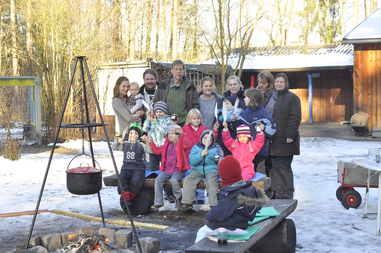 Eine Gruppe Kinder steht mit ihren Eltern und dem Leiter der Veranstaltung auf dem Hof des Abenteuerspielplatzes. Links hängt ein Topf an einem Gestell über einem Lagerfeuer.      