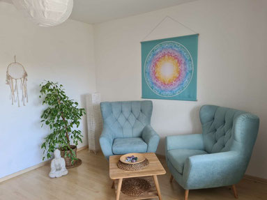 Gemütlicher Praxisraum mit zwei blauen Sesseln, einem kleinen Holztisch und einem Mandalabild an der Wand
