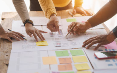 Mehrere Hände arbeiten gemeinsam an Strategien und Layouts auf einem Tisch – visuell als Symbol für Veränderungsprozesse in KMUs und Teams im Wandel.