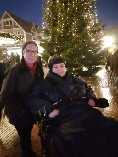 Johanna, im E-Rollstuhl. Links daneben steht ihre Mutter. Im Hintergrund ist ein großer beleuchteter Weihnachtsbaum und ein Weihnachtsmark. Beide lächeln in die Kamera. (2022)