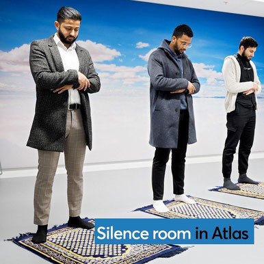 3 male students standing next to eachother in the silence room on small carpets with their arms over each other. They are standing in front of a wallpapered wall with a blue sky and white clouds.