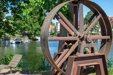 Am Ufer des Flusses Havel steht ein großes Rad aus rostigem Eisen. Im Hintergrund sieht man den Fluss, Bäume und Häuser.