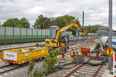 Auf dem Fernbahngleis Am Kombibahnsteig in Blankenfelde steht ein Zweiwegebagger mit Betonkübel, der gerade vom Fahrmischer befüllt worden ist.