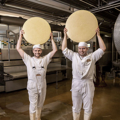 Zwei Personen in Hygienekleidung stehen im Produktionsbereich und halten Käse in den Händen