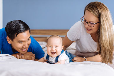 Ein Baby liegt zusammen mit den Eltern auf dem Elternbett und lacht in die Kamera.