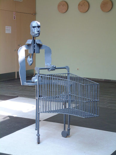 Shopping for Faces — early version of the Vintage Cyborg Shopping Cart sculpture featuring a metal cyborg figure pushing a shopping cart, exhibited at CC De Adelberg, showing a visible mechanical heart and industrial construction.