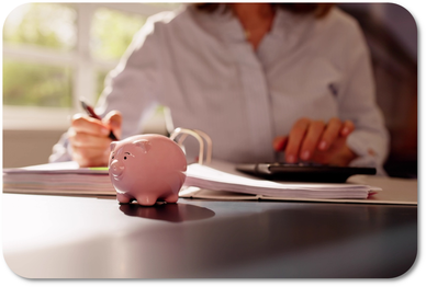 Eine Frau am Schreibtisch sitzend mit einem Kugelschreiber und Taschenrechner in der Hand. Im Vordergrund ein rosa Sparschwein.