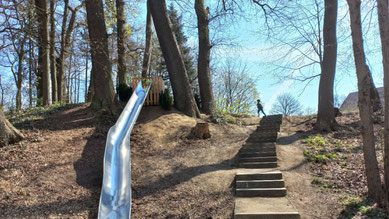 Große Rutsche und eine Steintreppe inmitten von großen Bäumen im Park, auf der obersten Treppe ist ein Kind zu erkennen