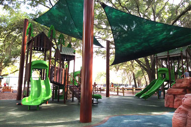 木々に囲まれたトロットウッド公園（Trotwood Park）の遊具エリア。緑色の滑り台やアスレチック遊具が設置され、上部には大きな日よけシェードが張られていて真夏でも安心。ウィンター・スプリングスの住宅街にあり、水遊びや筋トレエリア、池の散策も楽しめる家族向けの人気スポット。