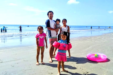 ココアビーチの海辺で家族が楽しむ様子。白い砂浜と青い海が広がり、子どもたちは水着や浮き輪を持って元気に遊び、父親が小さな子を抱えて記念撮影。オーランド中心部から車で約1時間で行ける手軽なビーチで、サーフィンや散策、観光客に人気の「ココアビーチ・ピア」などがある定番スポット。
