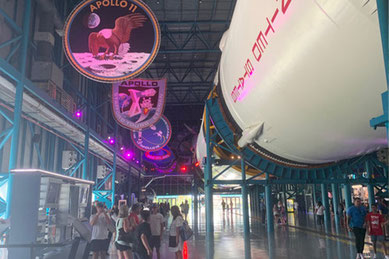 巨大なサターンVロケットが展示されるケネディスペースセンターの館内。天井からはアポロ計画のミッションパッチが吊り下げられ、多くの観光客がその迫力に見入っている。NASAやSpaceXの有人打ち上げ拠点として知られ、アポロ計画やスペースシャトル計画の歴史を体感できる宇宙開発の聖地。