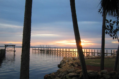 夕暮れのケープ・カナベラル海岸。ヤシの木の間から沈む夕日が海に反射し、静かな波と桟橋が美しく映える風景。フロリダ東海岸に位置するこの岬は、宇宙開発の拠点やクルーズ港、アウトドアの人気スポットとして知られ、ココアビーチからも近く日帰り観光に便利なアクセスが魅力。