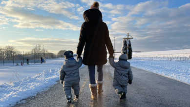 Mama läuft in Winterlandschaft auf der Straße, an jeder Hand hat sie einen kleinen Jungen. Alle sind warm eingepackt und von hinten zu sehen.