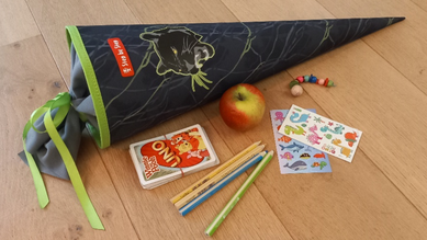 Eine Schultüte liegt auf dem Boden, daneben ein Apfel, ein Kartenspiel, Stifte, ein Schlüsselanhänger und Kindertattoos