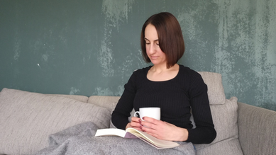 Michaela Dauscher sitzt auf dem Sofa und liest ein Buch, in ihrer Hand hält sie eine Tasse, sie ist zugedeckt, die Wand hinter ihr ist grün
